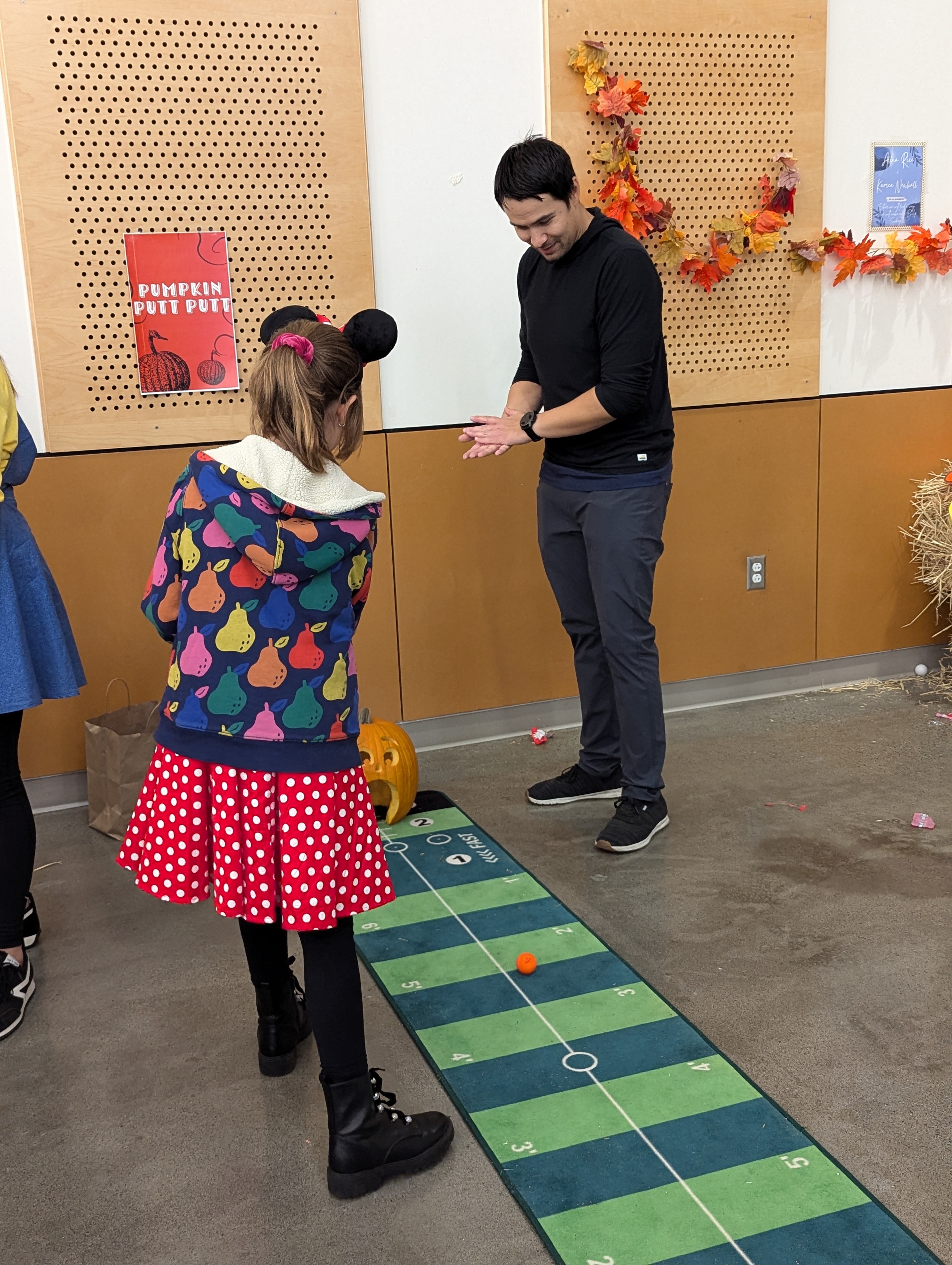 A student puts a golf ball into a jack-o-lantern's mouth while a parent volunteer cheers her on.